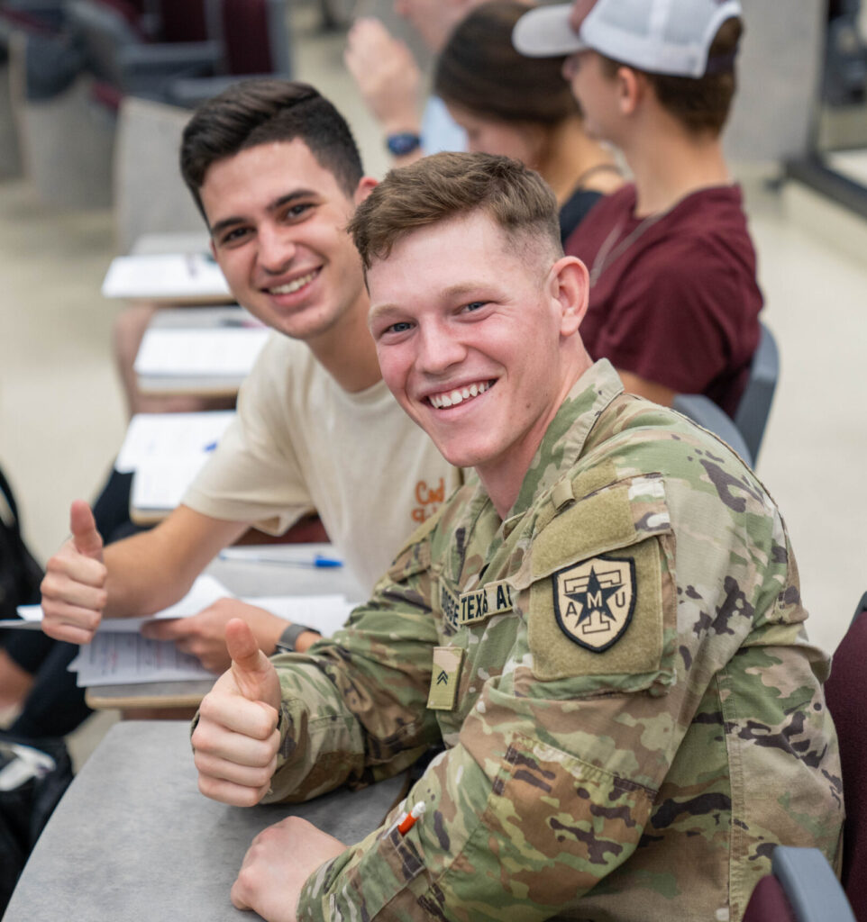 two students, one in uniform, sitting and giving a thumbs up