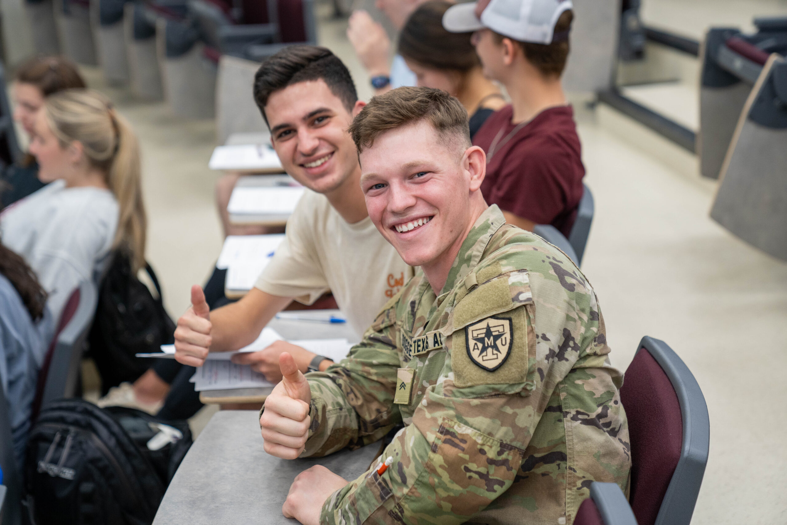 two students, one in uniform, sitting and giving a thumbs up